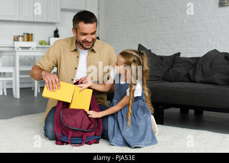 Padre sorridente aiutando piccola figlia imballaggio zaino per il primo giorno di scuola, torna al concetto di scuola Foto Stock