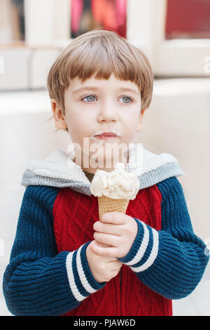 Closeup ritratto della cute bianca bionda caucasica ragazzo di età prescolare con occhi verdi in felpa rossa mangiare leccare il gelato in cono di cialda. Foto Stock