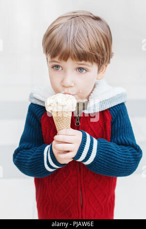 Closeup ritratto della cute bianca bionda caucasica ragazzo di età prescolare con occhi verdi in felpa rossa mangiare leccare il gelato in cono di cialda. Foto Stock