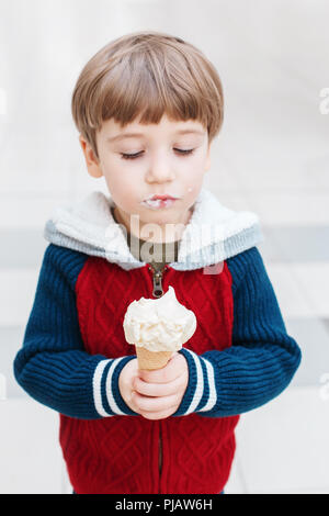 Closeup ritratto della cute bianca bionda caucasica ragazzo di età prescolare con occhi verdi in felpa rossa mangiare leccare il gelato in cono di cialda. Foto Stock