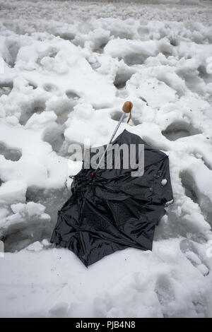 Close up rotto accartocciata Ombrello nero giacente scartato in scongelamento coperta di neve con impronte sul ciglio della strada Foto Stock