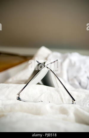 Close up gli occhiali da lettura appoggiati davanti a un libro in brossura aperto su un piumone bianco nel letto con camera da letto in background Foto Stock