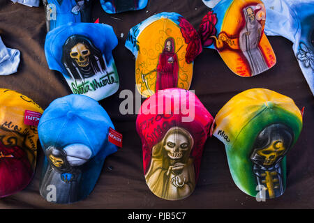 Colorato i cappelli da baseball e con vari Santa Muerte (Santa morte) foto, vengono venduti per strada durante un pellegrinaggio a Città del Messico. Foto Stock