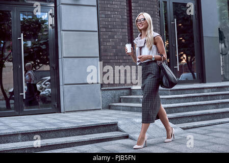 Femmina lavoratore officer di camminare sulla strada del centro cittadino con il caffè del mattino. Fiducioso imprenditrice in elegante usura formale guarda alle vetrine di negozi e caffè di contenimento per andare Foto Stock