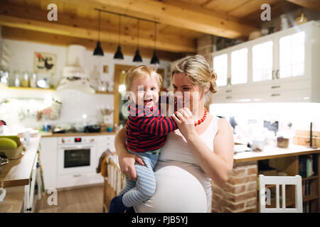 Bella donna incinta trasporta un bambino ragazzo in cucina a casa. Foto Stock