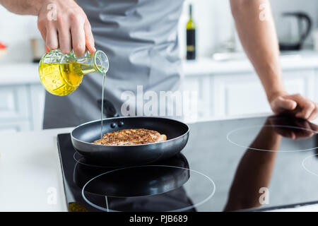 Ritagliato colpo di uomo in grembiule versando olio durante la cottura bistecca in padella Foto Stock