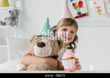 Sorridente compleanno kid holding Teddy bear e dando cupcake Foto Stock