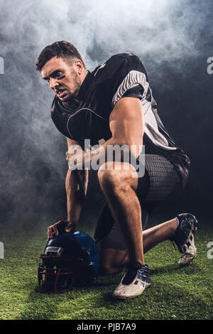 Arrabbiato giovane giocatore di football americano con il casco in piedi sul ginocchio su nero Foto Stock