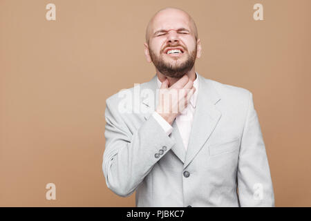 Gola del dolore o di freddo. ritratto di mezza età calvo imprenditore barbuto con gli occhi chiusi in grigio chiaro tuta in piedi e toccando la sua dolorosa collo. indo Foto Stock