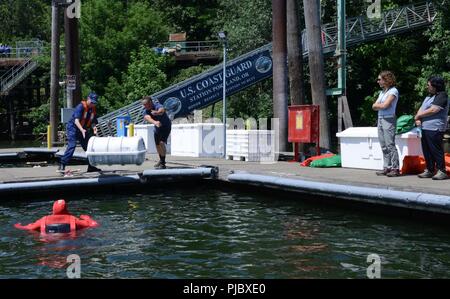 Ron Hilburger, Guardia costiera, ausiliario e il Tenente j. g. Adam Birch, nave statunitense ispettore di sicurezza marina Portland unità, dimostrare come distribuire una zattera gonfiabile canister per membri del Washington Homeland Security Tavola Rotonda nel fiume Willamette in bacini marini di unità di sicurezza Portland, Ore., 16 luglio 2018. Zattere di salvataggio sono uno dei tanti necessari elementi di sicurezza su tutti i dipendenti degli Stati Uniti e stranieri di navi commerciali. Stati Uniti Coast Guard Foto Stock