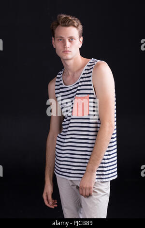 Bel giovane con gli occhi blu in una t-shirt casual con strisce e pantaloni bianchi su sfondo scuro Foto Stock
