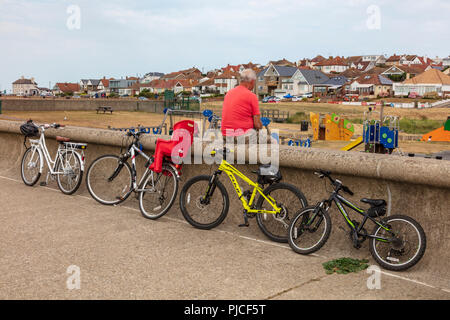 Un nonno siede sulla parete del mare a Hampton e guarda a sua moglie e i nipoti (invisibile) nel parco giochi, le loro biciclette appoggiarsi contro la parete. Kent, Regno Unito Foto Stock