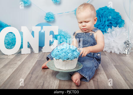 Ritratto di carino adorabile bionda Caucasian baby boy con gli occhi blu in jeans complessivamente, festeggia il suo primo compleanno con torta gourmet, lettere uno e Foto Stock