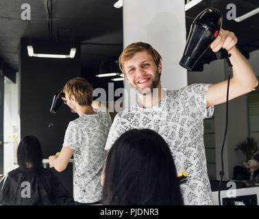 Professional sorridente stilista maschio soffiare l'asciugatura donna capelli con un essiccatore in salone Foto Stock