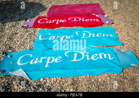 Tre coloratissimi asciugamani da spiaggia con la scritta "Carpe Diem", distesa su una spiaggia di ciottoli con rocce poste sui bordi per pesare giù dal vento Foto Stock