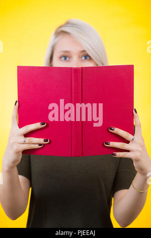 Giovane donna che copre metà faccia con una apertura libro rosso. Presentazione autore, mostrando la sua nuova creazione, focus sul libro isolato su giallo backgroun Foto Stock