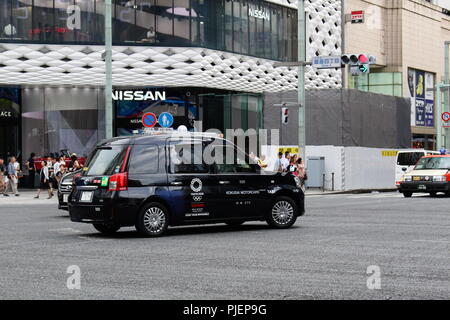 Toyota JPN Taxi con Tokyo logo olimpico su di esso passando attraverso una giunzione di Ginza davanti al Ginza luogo di costruzione. Una leggera sfocatura di movimento. Agosto 2018 Foto Stock