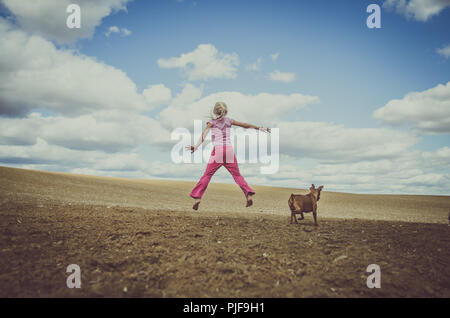 Bambina di salto e animale cane insieme in campo nella campagna sotto scenic cielo nuvoloso tono opaco Foto Stock