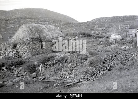 Degli anni Cinquanta, storico, tradizionale crofter's cottage in pietra con il tetto di paglia, situato sul pendio di una collina nella scarsa paesaggio rurale delle Ebridi Esterne, Western Isles Scotlamd, UK. Foto Stock