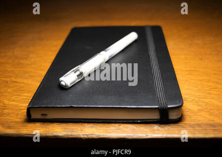 Penna bianca su un libro nero sulla parte superiore di una scrivania in legno. Strumenti di scrittura per un autore. Foto Stock