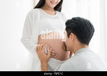 Asian marito baciare incinta pancia di sua moglie con gli occhi chiusi. Asian coppia sposata e il concetto di famiglia. Foto Stock