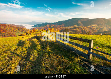 La bellissima campagna a foggy autunno sunrise. staccionata in legno attraverso le zone rurali di prato alpino chioma rossa sulle colline boscose. Il cloud inversione nel lontano v Foto Stock