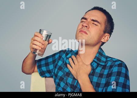 Un dolore terribile nella sua gola. Frustrati bel giovane toccando il suo collo e tenendo un bicchiere d'acqua. Sete con una sbornia Foto Stock