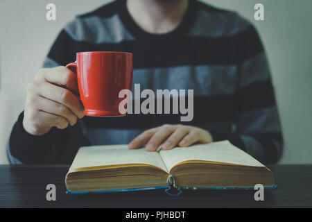 Uomo libro di lettura con caffè o tè. Rosso coppa in mano. Un uomo in un maglione a righe. Foto Stock