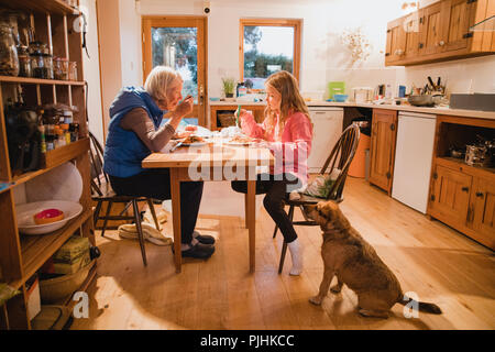 Vista laterale di una nonna e la nipote seduti al tavolo da pranzo. Essi sono entrambi di mangiare il loro pasto. Sul pavimento accanto alla bambina th Foto Stock