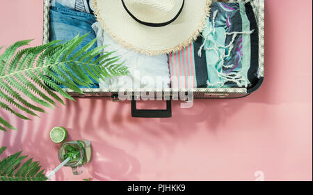 Foglia di felce, tropicale acqua detox e aprire la valigia con vestiti sul rosa pastello. Vista da sopra con copia spazio. Concetto di estate viaggio. Foto Stock