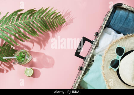 Foglia di felce, tropicale acqua detox e aprire la valigia con vestiti sul rosa pastello. Vista da sopra con copia spazio. Concetto di estate viaggio. Foto Stock