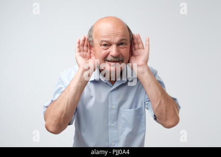 Ritratto di senior uomo informale che overhears conversazione su sfondo bianco. Parlare ad alta voce si prega di concetto. Che cosa hai detto Foto Stock