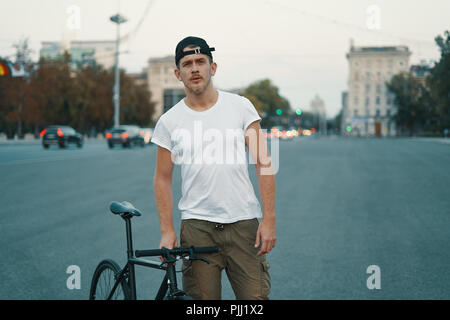 Outdoor ritratto di un moderno e giovane uomo della strada, e discosto bike. Un giovane uomo di atletica che indossa i pantaloni khaki, T-shirt bianco, cercando di fotocamera. Foto Stock