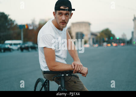 Outdoor ritratto di un moderno e giovane uomo della strada, seduto sulla bici. Un giovane uomo di atletica che indossa i pantaloni khaki, T-shirt bianco, cercando di fotocamera. Foto Stock
