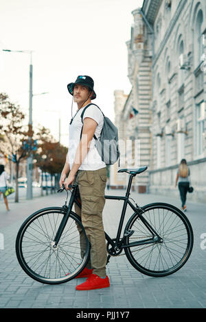 Outdoor ritratto di un moderno e giovane uomo della strada, seduto sulla bici. Un giovane uomo di atletica che indossa i pantaloni khaki, T-shirt bianco, rosso sneakers e backp Foto Stock