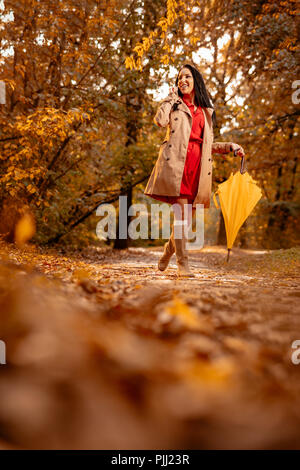 Felice giovane donna in abito rosso parlando sullo smartphone camminando in autunno soleggiato parco, tenendo ripiegati Ombrello giallo. Foto Stock