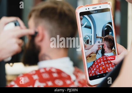 Un bel giovane uomo barbuto in un barbiere. Il barbiere spazzole i suoi capelli e lo taglia con un rifinitore elettrico. Foto Stock