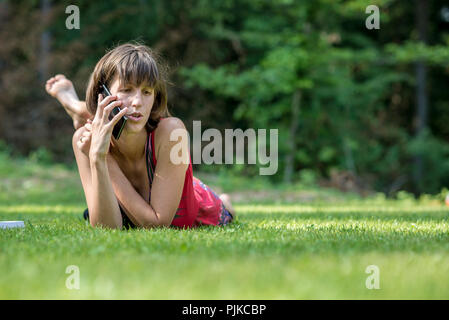 Giovani brunette giacente in erba verde appoggiata sulle sue braccia come parla al telefono cellulare. Foto Stock