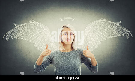 Entusiasta giovane donna dando una doppia come Thumbs up gesto immaginare se stessa come un angelo con le ali dietro la schiena e un alogeno sopra la testa. Super Foto Stock