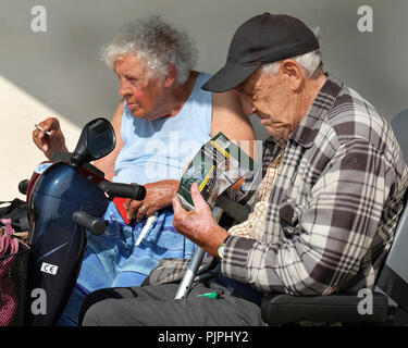 Il vecchio uomo e donna in scooter non valido di fumare all'esterno nella luce del sole. Foto Stock
