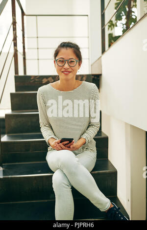 Sorridente giovane imprenditrice asiatici indossando occhiali da sole seduti sulle scale in un ufficio moderno mantenendo il suo cellulare Foto Stock