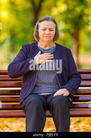 Senior lady stringendo il suo torace nel dolore ai primi segni di angina o un infarto del miocardio o un attacco di cuore, corpo superiore nel parco su una giornata autunnale Foto Stock