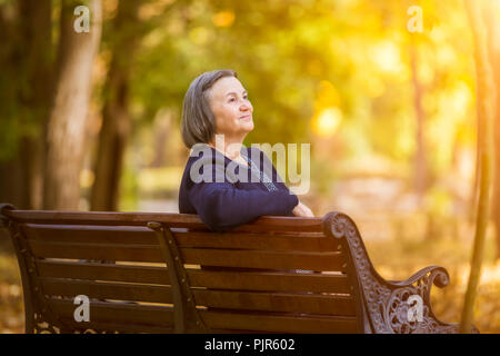 Donna anziana seduta su una panchina nel parco d'autunno. Età donna seduta su un banco di lavoro in autunno park guarda e sorridente al sun. Foto Stock