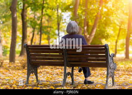 Donna anziana seduta su una panchina nel parco d'autunno. Lonely di età donna seduta su un banco di lavoro in autunno park guarda gli alberi. Foto Stock