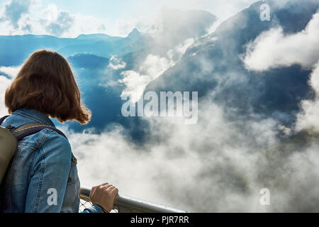 Una giovane donna con uno zaino in piedi dalla ringhiera oltre le montagne con le nuvole e la nebbia guardando giù nella valle di Hallstatt. Austria in Ue Foto Stock