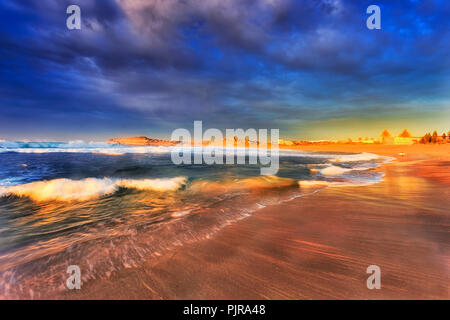 La sabbia lavata di Mona Vale beach in morbido caldo sole del mattino con le onde di laminazione ideale per praticare il windsurf sotto colorato cielo nuvoloso. Foto Stock