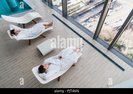 Due giovani uomo indossare accappatoio, sdraiato sul lettino in spa e salone di parlarsi di fronte ad una grande finestra con una fantastica vista della città. Spa concept Foto Stock