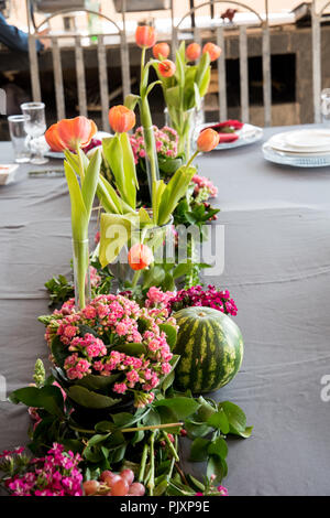 Un allestimento floreale di bellissimi fiori arancione con colorati di rosa mazzetto in primo piano con un invitante round anguria e uva. Foto Stock