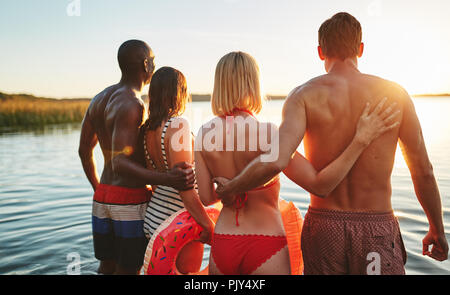 Specchietto di due coppie giovani in costumi da bagno e guardare il tramonto insieme mentre in piedi a braccetto in un lago Foto Stock