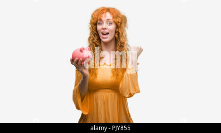 Giovane donna redhead holding salvadanaio urlando orgoglioso e celebrando la vittoria e il successo molto eccitato, allietarla emozione Foto Stock